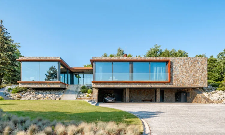 Dom na Skarpie, zdjęcie od strony podjazdu. Dom zaprojektowano jako parterowy, osadzony w zboczu, z częścią techniczną w przyziemiu. Widoczne duże szyby w dziennej części domu oraz elewacja z kamienia oświetlona promieniami słońca.